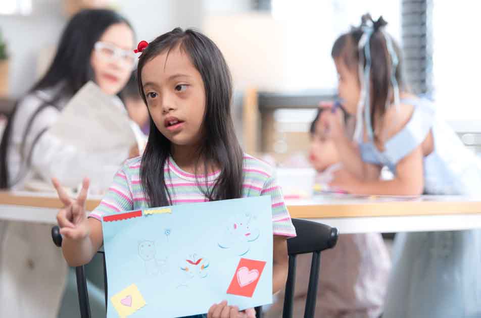 Young girl with Down syndrome proudly showing her artwork—representing expressive language growth through speech therapy in Phnom Penh.