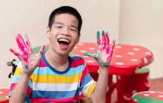 Smiling child with disabilities showing painted hands during inclusive art therapy session at OrbRom Center in Phnom Penh.