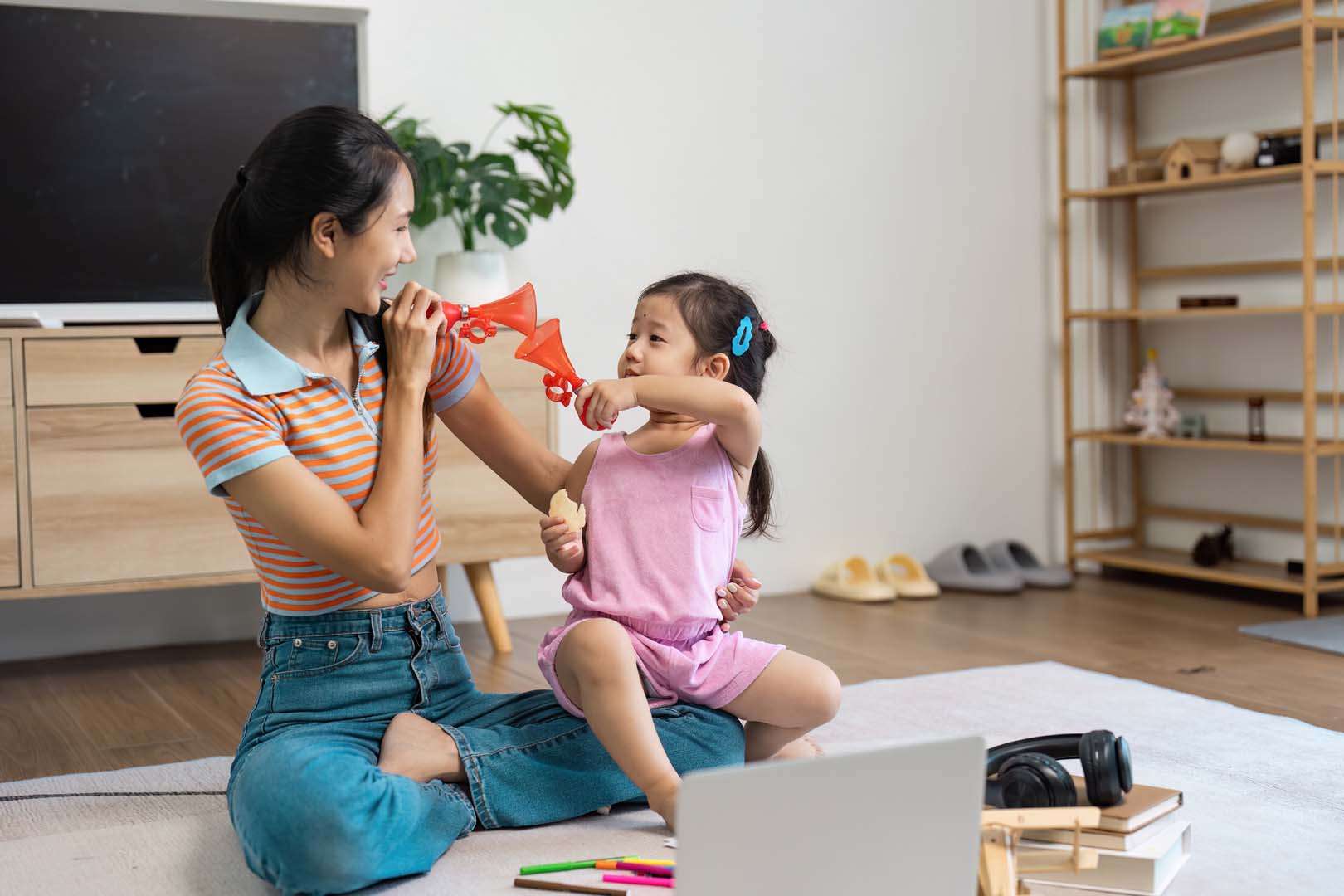 A smiling woman and young girl playing with red horn toys—demonstrating how sound play supports early speech and communication development.