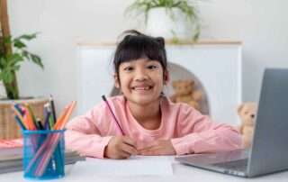 Smiling girl studying at a desk with laptop and pencils, representing confidence and joyful learning for children with learning differences.