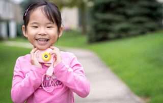 Young girl smiling and holding a toy camera, engaging in outdoor creative play and joyful self-expression