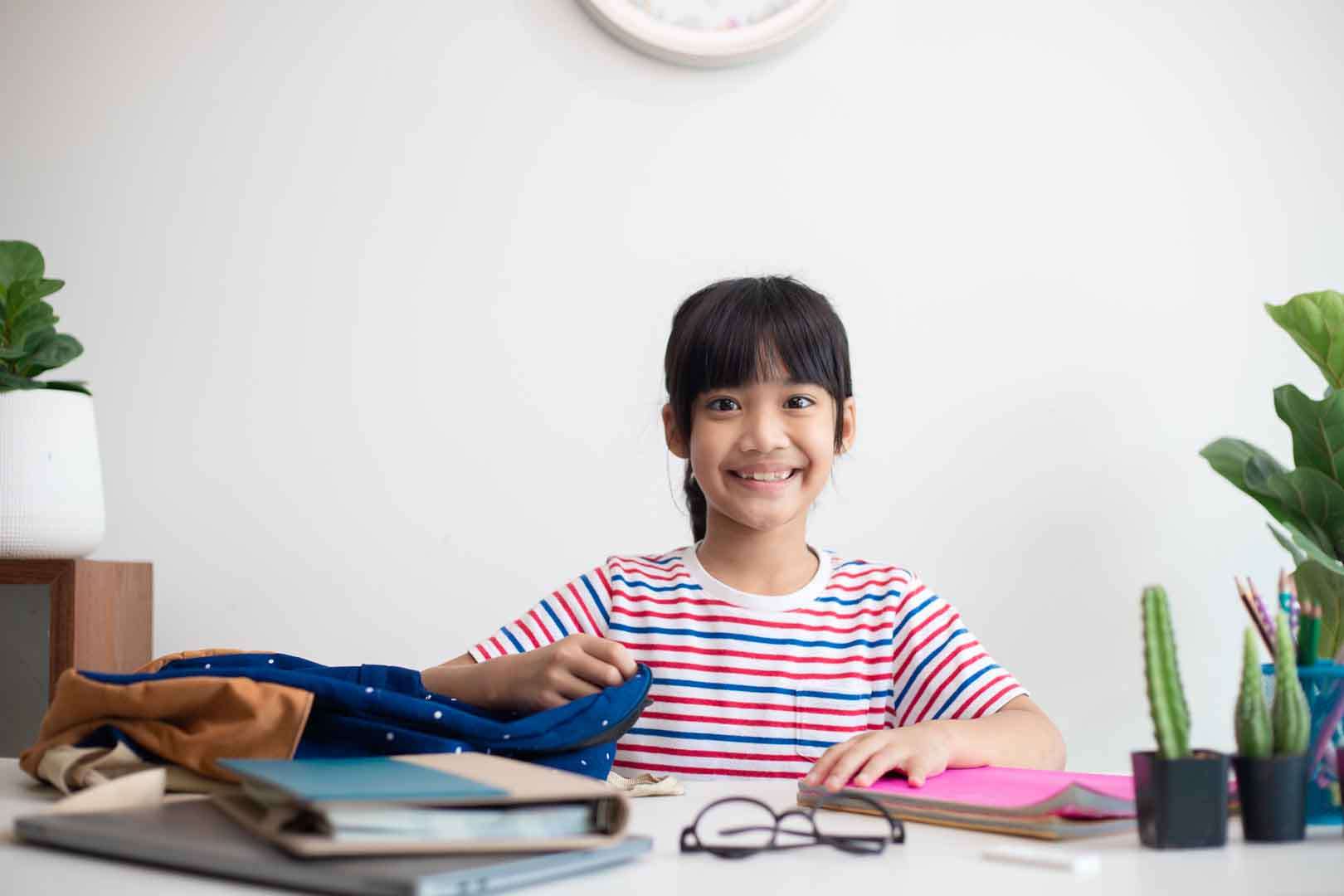 A young girl smiling at her school desk, ready for her day with organized school supplies – symbolizing independence and readiness.