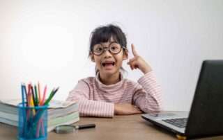Young girl wearing glasses with a bright expression, sitting at a desk with school supplies and a laptop, symbolizing gifted learning at home