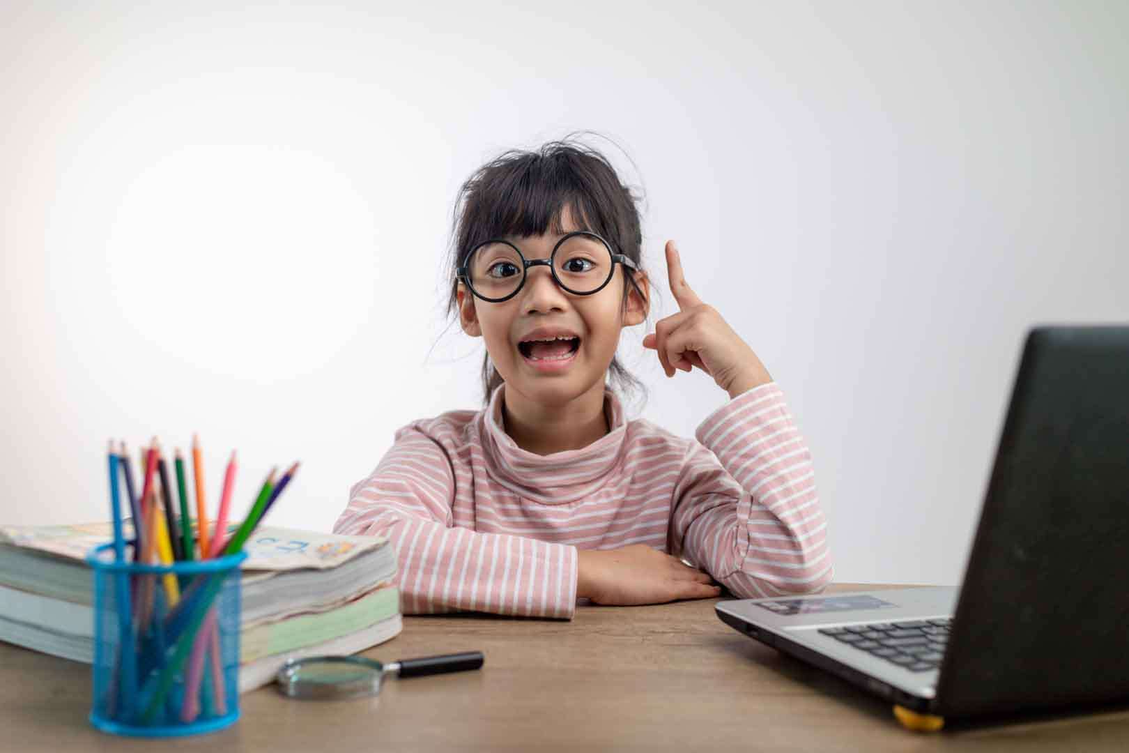 Young girl wearing glasses with a bright expression, sitting at a desk with school supplies and a laptop, symbolizing gifted learning at home