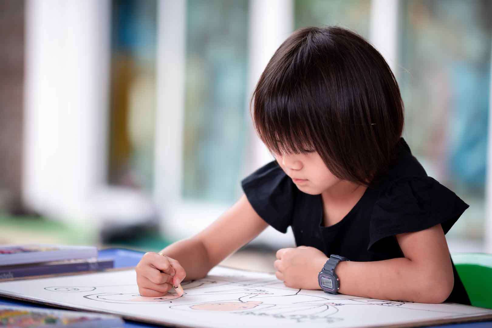 Young girl intently drawing with a crayon on a large sheet of paper during a fine motor skill activity